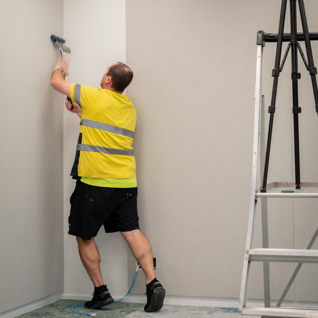 Person in yellow safety vest using a tool on a wall next to a tripod and ladder.